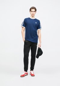 Tmavomodré tričko s bielym lemovaním a tromi bielymi pruhmi na rukávoch, čierne tepláky, červeno-biele tenisky, drží čiernu čiapku.