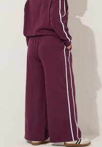 Weite Bein-Hosen aus tiefburgunderfarbenem Stoff mit weißen Seitenstreifen. Sie verfügen über einen elastischen Bund und eine lockere Passform, kombiniert mit beigen Sneakers.