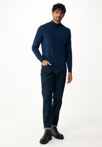 Suéter de cuello alto en algodón azul marino, de punto liso, combinado con jeans de mezclilla oscuros. El modelo lleva botas negras de tobillo con suela texturizada.