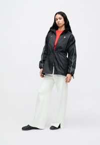 Μαύρο ελαφρύ μπουφάν με λευκές λεπτομέρειες, ελαστική μέση και κουκούλα, συνδυασμένο με κόκκινη μπλούζα και λευκό παντελόνι φαρδιάς γραμμής.