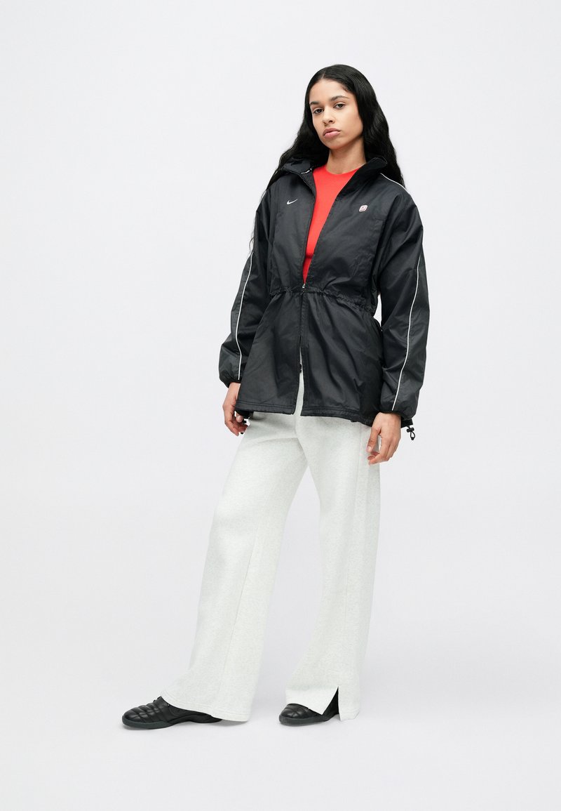 Schwarze leichte Jacke mit weißen Akzenten, elastischem Taillenbund und Kapuze, kombiniert mit einem roten Oberteil und weißen Hosen mit weitem Bein.