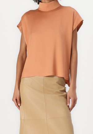 Femme portant un haut sans manches à col haut couleur pêche associé à une jupe beige longueur genou, debout avec les bras détendus.