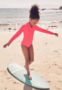 Felroze zwemkleding met lange mouwen en een ritssluiting aan de voorkant, gedragen door een kind dat balanceert op een lichtblauwe surfplank op een zandstrand, met de zonverlichte oceaan op de achtergrond.