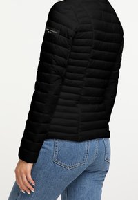 Person wearing a fitted black quilted jacket with a brand patch on the sleeve, paired with light blue denim jeans, shown from the back.