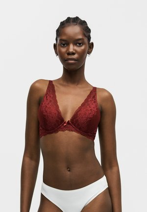 Donna con capelli intrecciati indossa un reggiseno di pizzo rosso scuro e slip bianchi, in piedi su uno sfondo grigio chiaro uniforme, rivolta verso di sé.