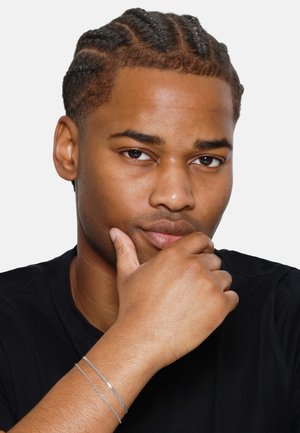 Bracelet en argent porté au poignet, présentant un design simple avec une texture lisse et élégante. Gros plan d'un visage avec des cheveux tressés et une expression pensive.