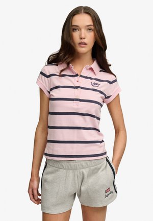 Młoda kobieta w różowej i granatowej paskowanej koszulce polo oraz jasnoszortach, stojąca z jedną ręką w kieszeni na białym tle.