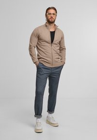 Maglione beige con zip e polsini a coste, abbinato a pantaloni grigi a gamba affusolata e sneakers bianche. Design minimalista con una silhouette elegante.