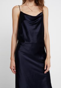 Robe nuisette en satin bleu marine à bretelles spaghetti, avec un col bénitier et une texture lisse, tombant jusqu'au milieu du mollet avec une silhouette ajustée.