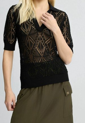 Femme portant un haut en dentelle noire à manches courtes avec des motifs géométriques et une jupe vert olive avec une poche et un rabat.