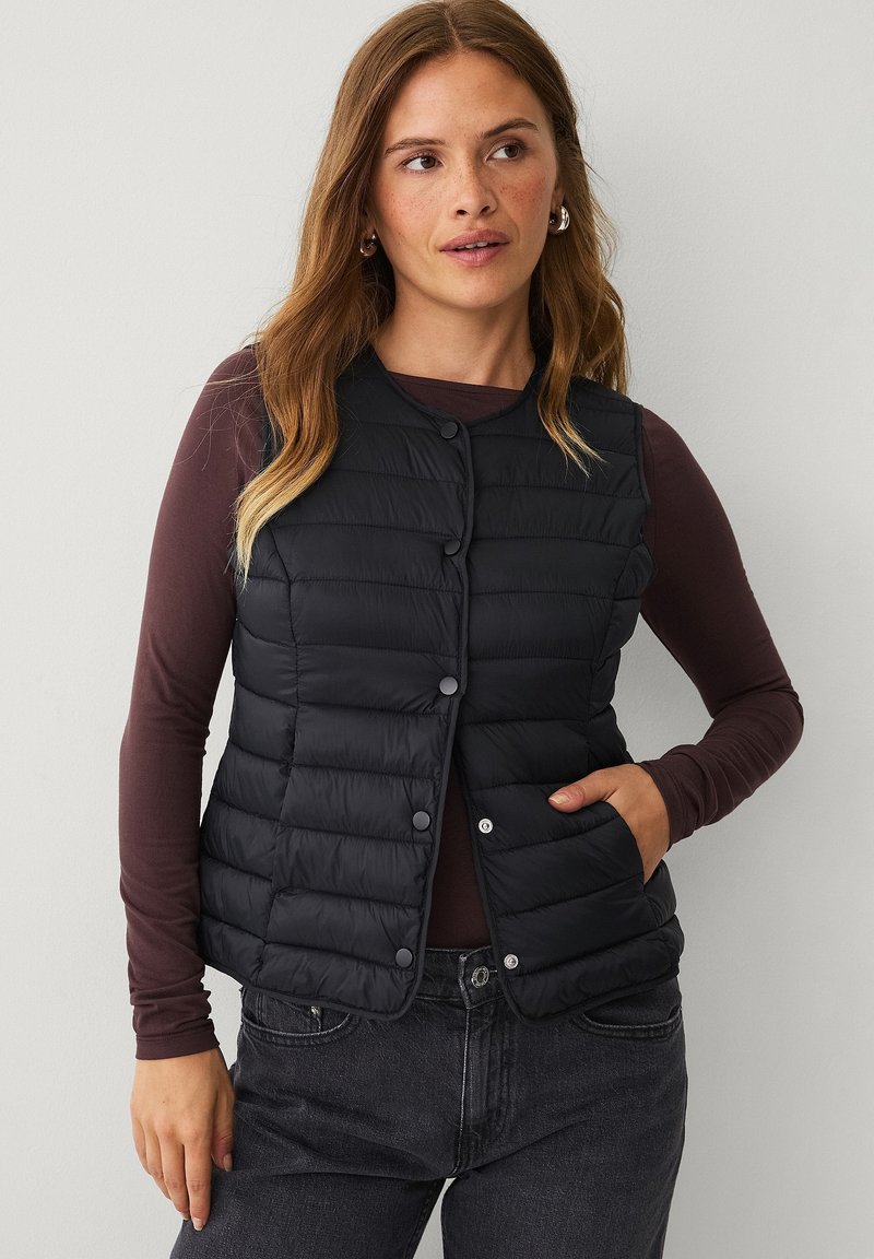 Schwarze Steppweste mit rundem Ausschnitt, Druckknöpfen und geripptem Texture, getragen über einem langärmeligen dunklen Oberteil mit seitlichen Taschen.