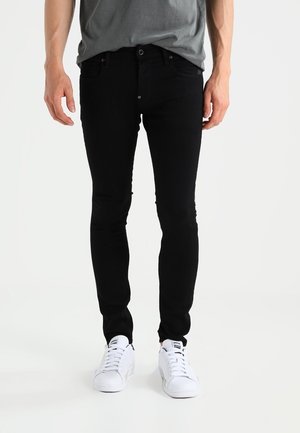 Homme portant un jean noir slim, un t-shirt gris et des baskets blanches, debout devant un fond uni.