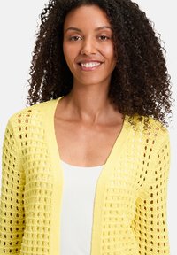Cardigan en tricot jaune avec un motif en maille ajourée et un décolleté en V, superposé sur un haut blanc, avec une finition douce et texturée.