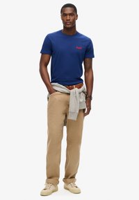 Camiseta de algodón azul marino con un logo rojo, combinada con pantalones beiges y una sudadera gris atada a la cintura; unos zapatos crema completan el conjunto.