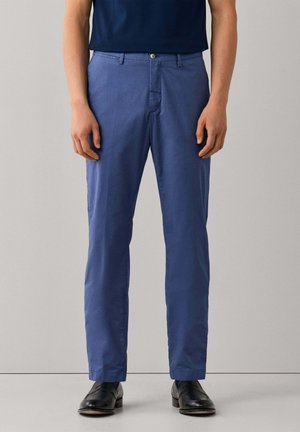 Man die op maat gemaakte blauwe broek draagt met riemlussen en zwarte nette schoenen, staand tegen een effen grijze achtergrond.