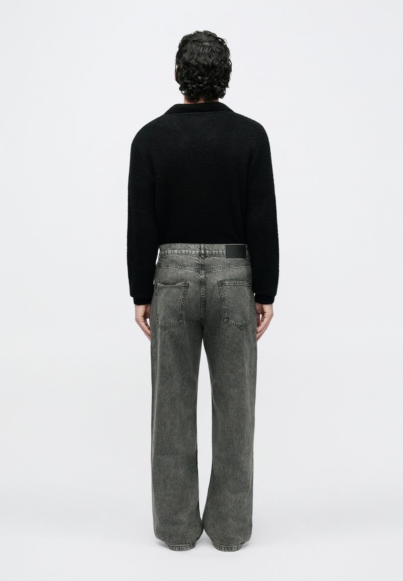 Man with curly dark hair wearing black sweater and gray wide-leg jeans standing facing a plain white background.