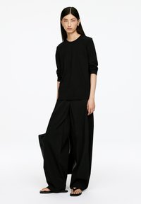 Schwarzes Langarm-T-Shirt mit lockerem Schnitt, kombiniert mit weiten schwarzen Hosen. Glatter Stoff mit minimalen Details. Getragen mit flachen Sandalen und einer Tragetasche.