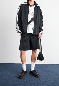 Svart idrottsjacka med vita ränder, svarta shorts, svarta skor och vita strumpor. Grafisk tröja med geometrisk design. Svart accessoarväska.