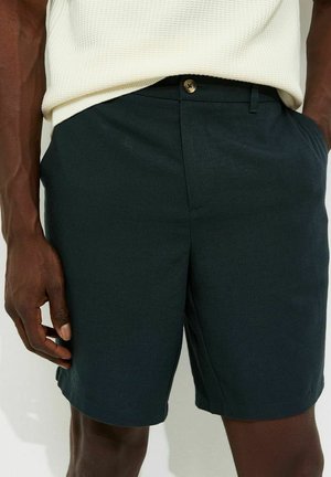 Shorts vert foncé ajustés avec bouton avant et poches, portés par une personne portant un haut clair texturé rentré.