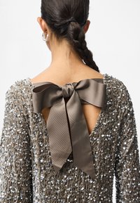 Φόρεμα με παγιέτες και βαθύ ντεκολτέ στην πλάτη, διακοσμημένο με μία μεγάλη, καφέ σατέν κορδέλα. Το ύφασμα έχει υφή, ενισχύοντας την οπτική πολυπλοκότητα του κομματιού.