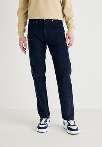 Pantaloni in corduroy blu scuro con un design a gamba dritta e una trama sottile, abbinati a sneakers bianche e navy, su uno sfondo neutro.