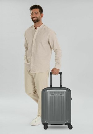 Mann in beige Outfit, lächelnd, steht mit der Hand in der Tasche und hält einen grauen SMARTBOX-Koffer am ausgezogenen Griff.