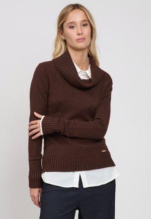 Junge Frau mit langen blonden Haaren, die einen braunen Rollkragenpullover, eine weiße Bluse und dunkle Hosen trägt und mit verschränktem Arm steht.