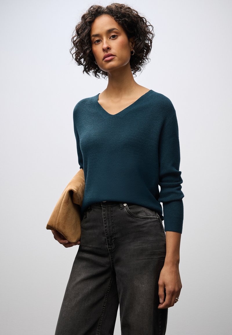 Teal-farbenes V-Ausschnitt-Pullover mit strukturiertem Strick, kombiniert mit hochgeschnittenen, dunkelgrauen Jeans. Hält eine hellbraune Clutch mit filigranen Details.