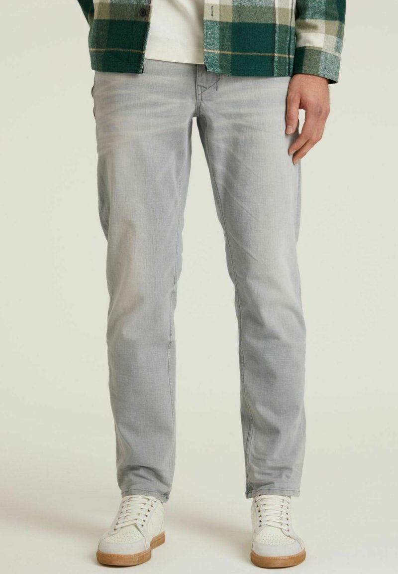 Vaqueros de un gris claro con un corte ajustado, que presentan un ligero desgaste. Combinados con zapatillas blancas y una camisa de cuadros verde.