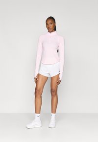 Top lung cu mânecă lungă de culoare roz deschis, cu guler cu fermoar pe jumătate, asociat cu pantaloni scurți albi. Modelul poartă adidași albi și șosete până la glezne, branding minim vizibil.