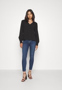Blusa nera a maniche lunghe con scollatura a V e dettagli arricciati, abbinata a jeans blu aderenti e tacchi neri con cinturini.
