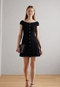 Robe noire plissée à manches courtes, avec un décolleté arrondi et des boutons argentés sur le devant. Accompagnée d'un petit sac noir.