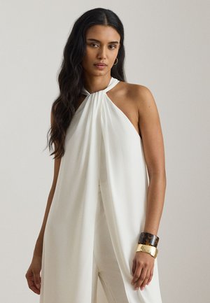 Kobieta o długich ciemnych włosach nosi białą, bezrękawną kombinezon z odkrytymi ramionami, złote kolczyki koła oraz masywne złote i żółwiowe bransoletki na prawym nadgarstku.