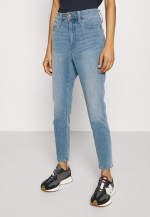 Henkilö, jolla on ylipolven korkealle nousevat vaalean siniset farkut ja mustavalkoiset sekä marjapuuro-väriset lenkkarit, yksinkertaisella vaalealla taustalla.