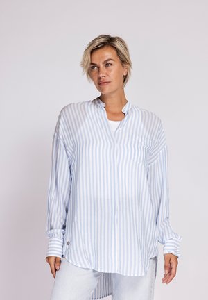 Frau mit kurzen blonden Haaren, die ein locker sitzendes, langärmliges, hellblau-weiß gestreiftes Hemd und helle Hose trägt und zur Seite schaut.