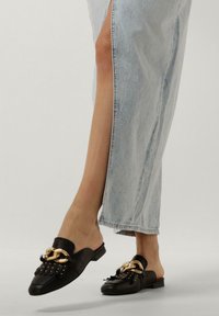 Zwarte leren muilen met een gouden kettingaccent, voorzien van franjes en zilveren studs, gecombineerd met een lichtblauwe denimrok.