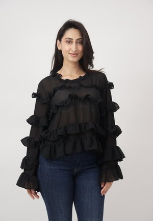 Frau trägt eine durchscheinende schwarze Bluse mit mehrlagigen Rüschen und blaue Jeans, steht vor einem schlichten hellen Hintergrund.