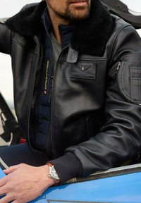 Veste bomber en cuir noir avec un col en fourrure, portée par-dessus un gilet matelassé bleu ; la montre-bracelet présente un cadran argenté et un bracelet en cuir marron.