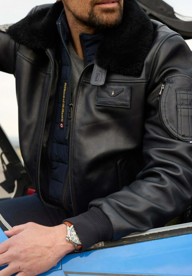 Veste bomber en cuir noir avec un col en fourrure, portée par-dessus un gilet matelassé bleu ; la montre-bracelet présente un cadran argenté et un bracelet en cuir marron.