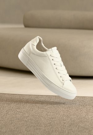 Witte leren sneaker met veters die boven een gestructureerd beige tapijt zweeft, met een beige bank op de onscherpe achtergrond.