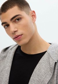 Joven con cabello corto castaño que lleva una camiseta negra y un cárdigan gris, mirando ligeramente hacia la cámara frente a un fondo sencillo.