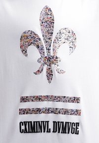 Bílé tričko s velkým, vícebarevným motivem fleur-de-lis a dvěma vodorovnými pruhy, doplněné výrazným černým textem pod nimi.