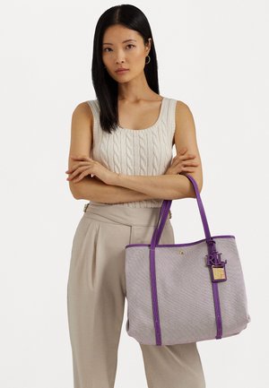 Mujer con cabello largo y negro que lleva un top de punto color crema sin mangas y pantalones beige, sosteniendo un bolso tote beige con asas y detalles morados.