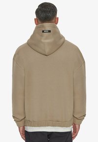 Hanorac oversized bej, cu glugă ajustabilă, manșete și tiv cu nervuri. Dispune de un mic etichetă logo neagră la spate. Textură de țesătură netedă.