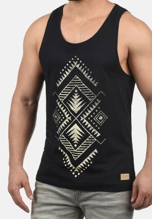 Homme portant un débardeur noir avec un motif géométrique tribal blanc sur le devant et un jean gris clair.