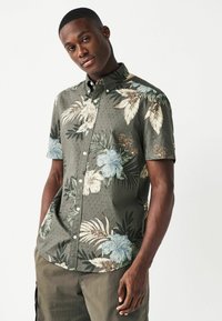 Camisa de manga corta con botones en tela verde oscura con estampados florales en azul, beige y verde. Superficie texturizada con detalles sutiles.