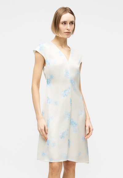 Γυναίκα που φοράει άσπρο αμάνικο φόρεμα μέχρι το γόνατο με ανοιχτό μπλε λουλουδάτα σχέδια, στέκεται μπροστά σε ένα απλό φωτεινό φόντο.