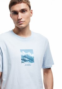 Joven con cabello corto que lleva una camiseta de color azul claro con un gráfico de olas del océano y el texto "Navega hacia nuevos horizontes."