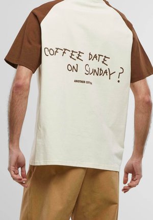 Mężczyzna noszący beżową i brązową koszulę z krótkim rękawem z tekstem "Coffee date on Sunday?" i "Another Cotton" na plecach, w połączeniu z brązowymi spodniami.