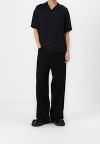 Camisa negra de manga corta y corte relajado, con botones en la parte delantera. Combinada con pantalones negros de corte holgado y zapatos negros de suela gruesa.